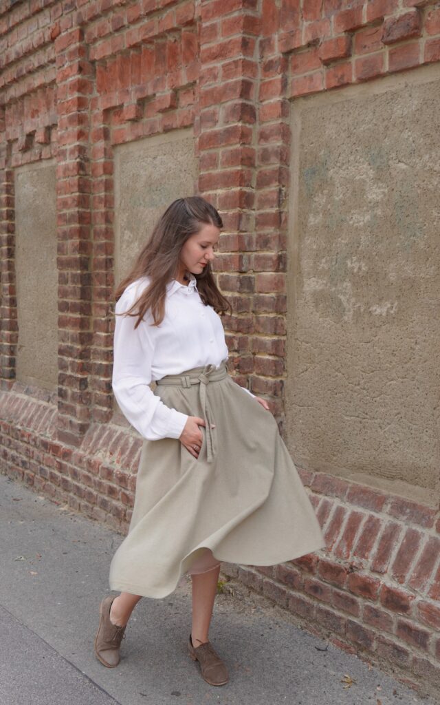 Young woman wears a light academia trend inspired outfit: a beige woolen Vintage skirt and a white vintage blouse. She enjoys to rewear Vintage , her flowy skirt moves in the wind.